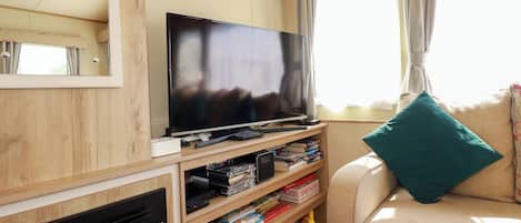 Una habitación con una televisión sobre un soporte de madera, una estantería con libros y juegos, un sofá con un cojín azul turquesa y una ventana con cortinas.