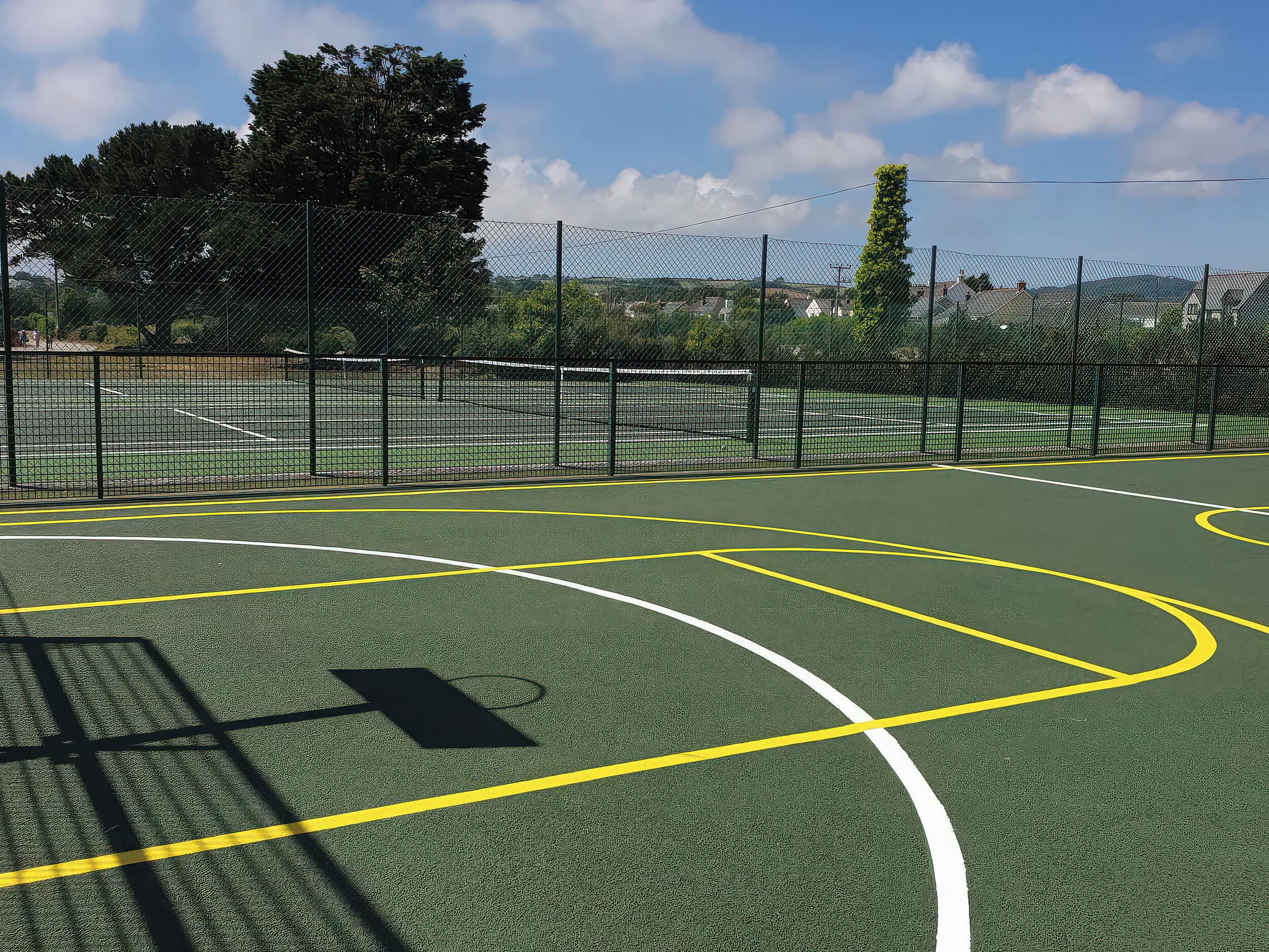 A basketball court with a chain-link fence, white lines marking the court, and a shadow of a basketball hoop.