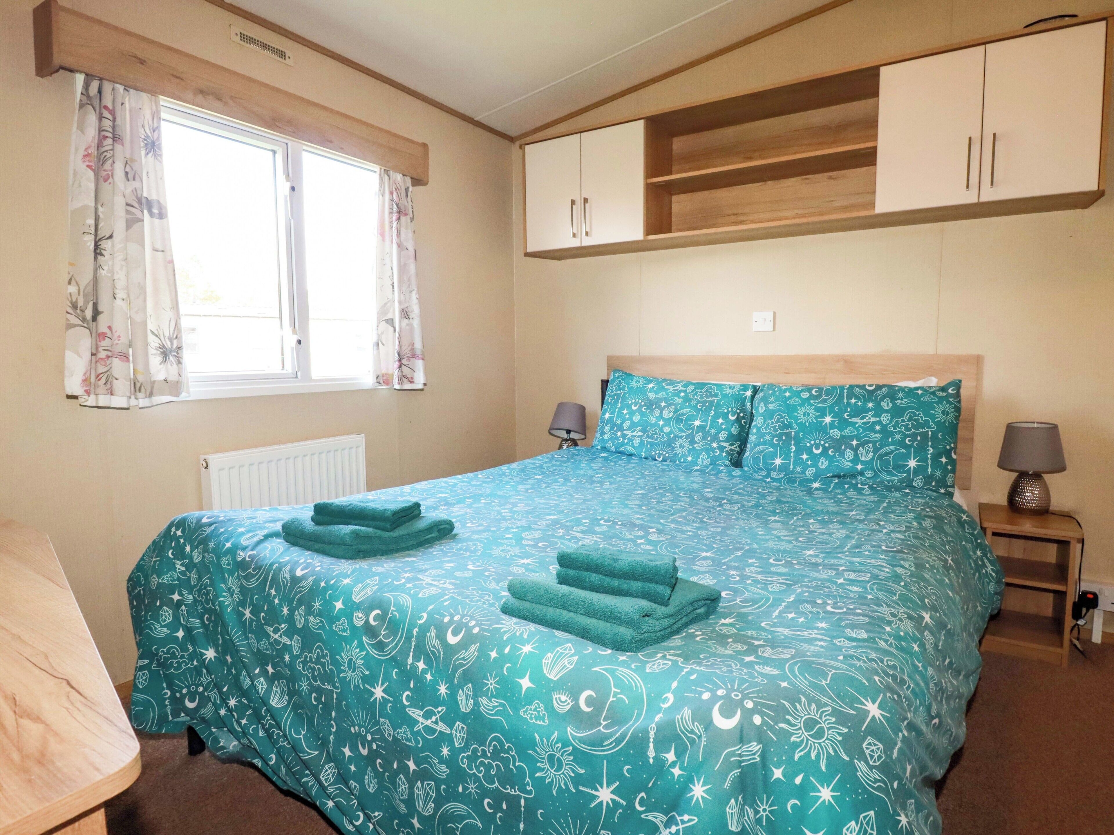 A bedroom with a bed, two bedside tables, a window with curtains, and built-in wooden cabinets above the bed.