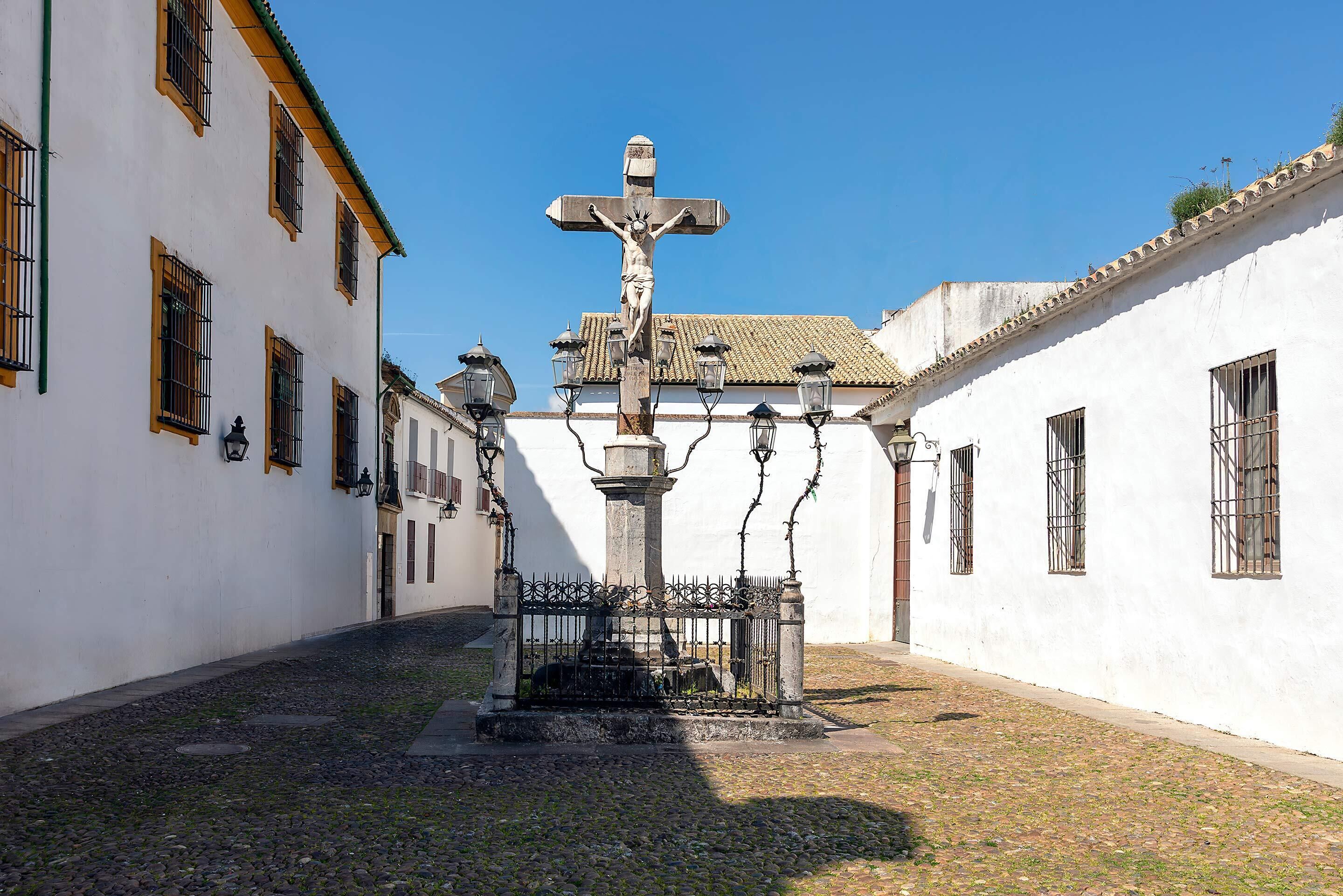 A narrow street with a crucifix at the end, white buildings with barred windows, and a cobblestone path.