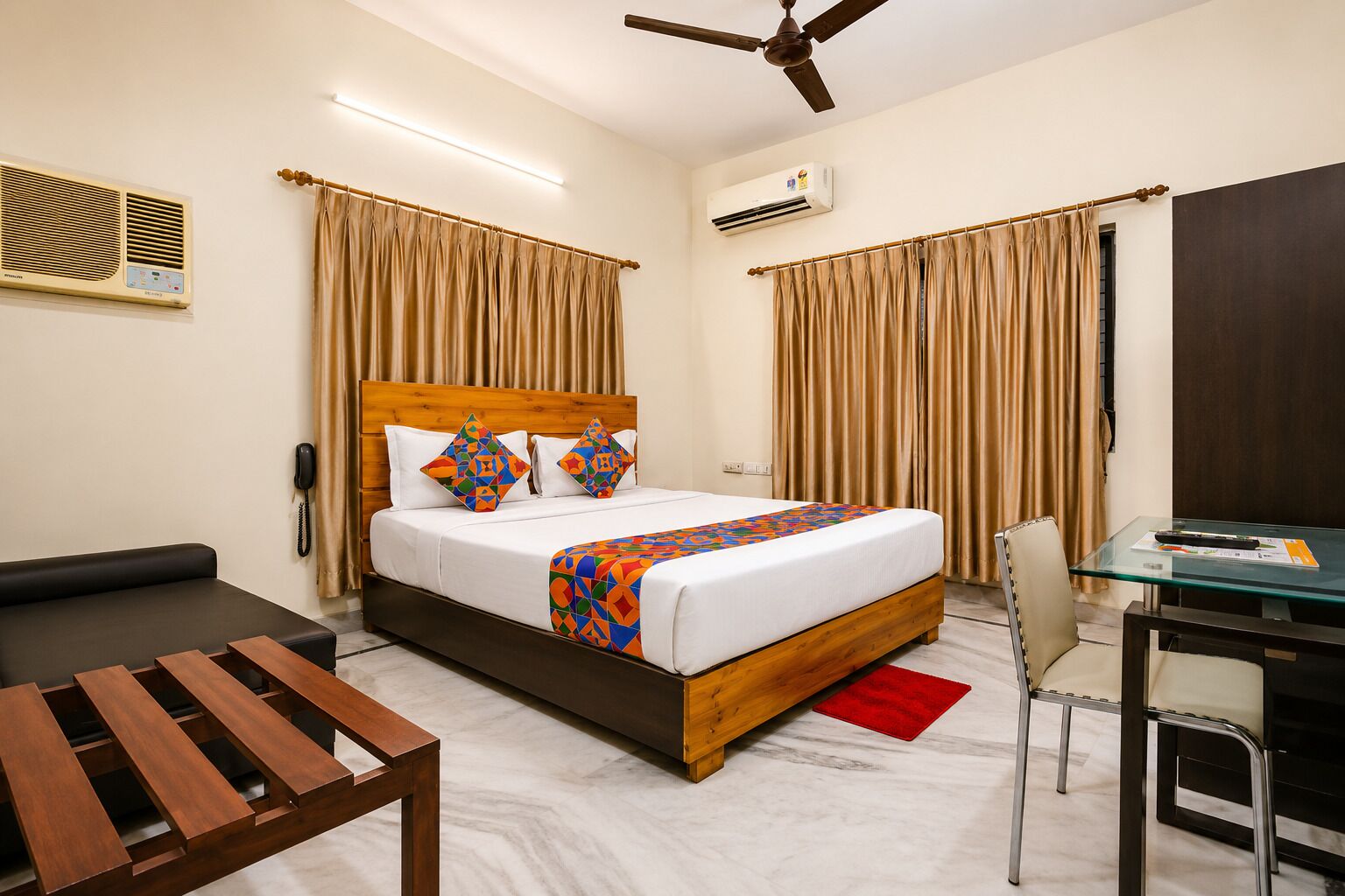 A hotel room with a bed, a desk, a chair, a wooden table, curtains, an air conditioning unit, and a ceiling fan.