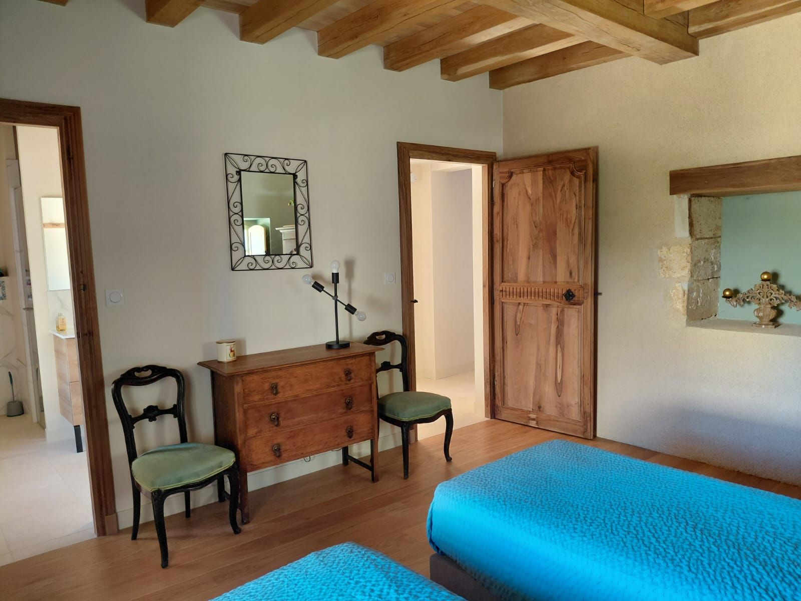 A room with a wooden dresser, two chairs, a bed with blue bedding, a mirror, and a wooden door.