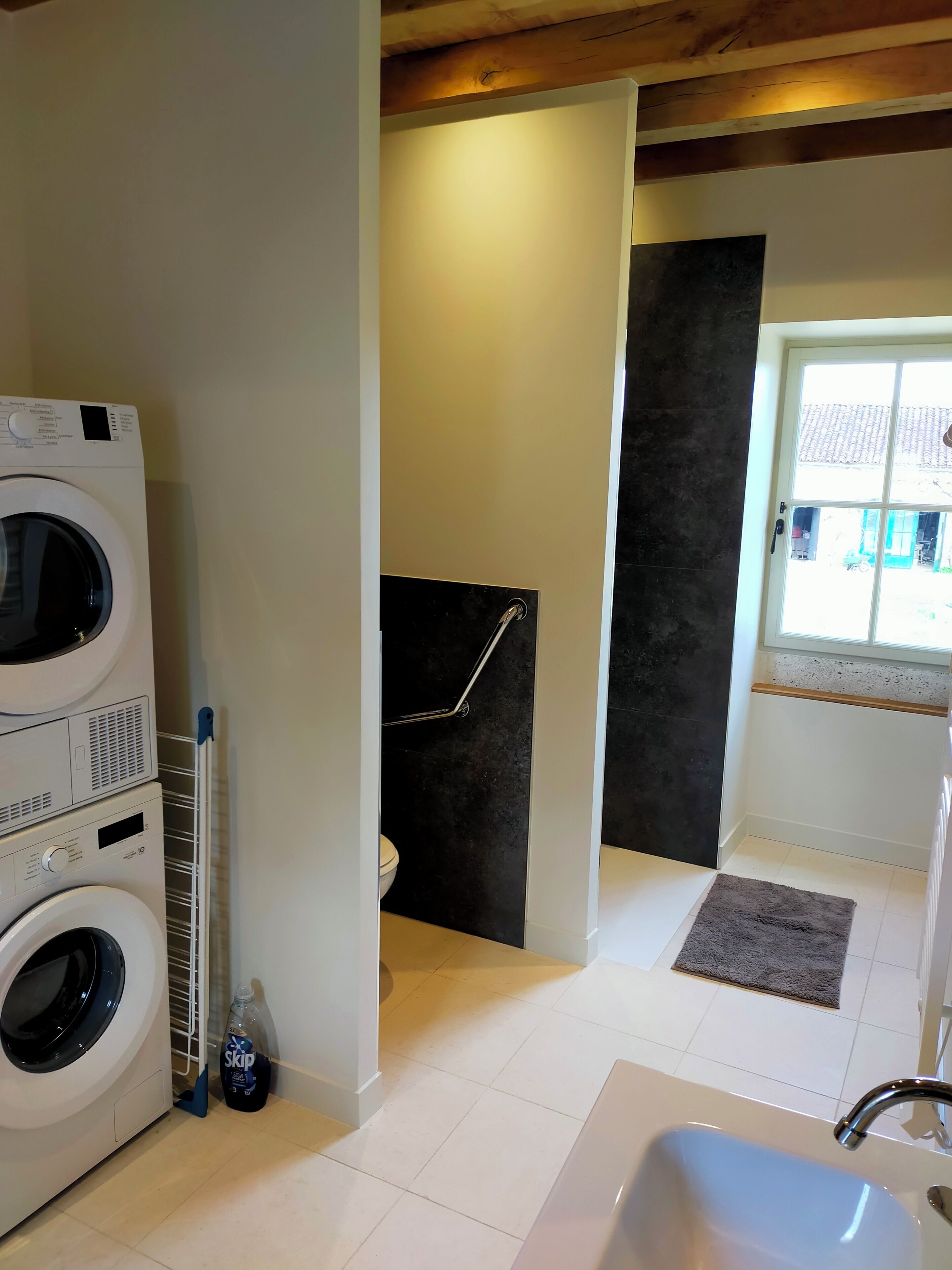 A laundry area with stacked washing machines, a shower with a black wall, and a window with a view of the outside.