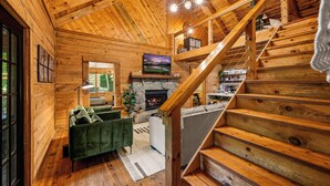 A wooden cabin interior with a fireplace, a green sofa, a staircase, and a television. - Gimme the Smokies, 2 BR, 2 BA, Sleeps 6 (Townsend)