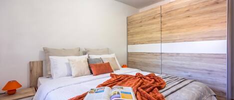 A modern bedroom with a wooden headboard, a bed with white bedding, multiple pillows, a red blanket, and a book on the bed.