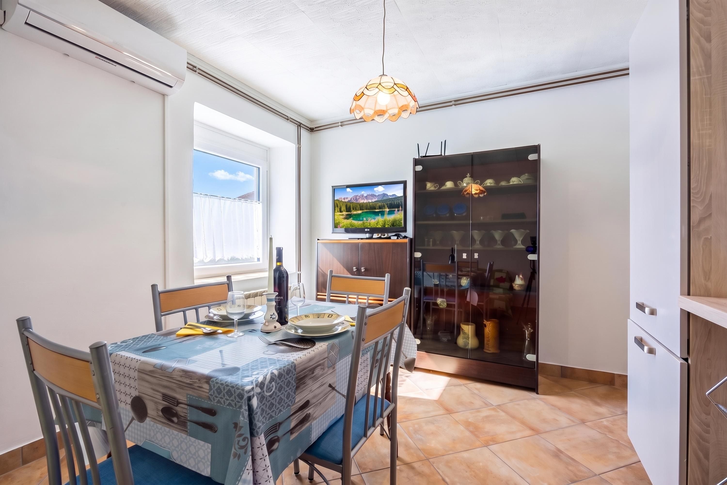 A dining area with a table set for two, a television mounted on the wall, and a cabinet with glass doors containing kitchenware.