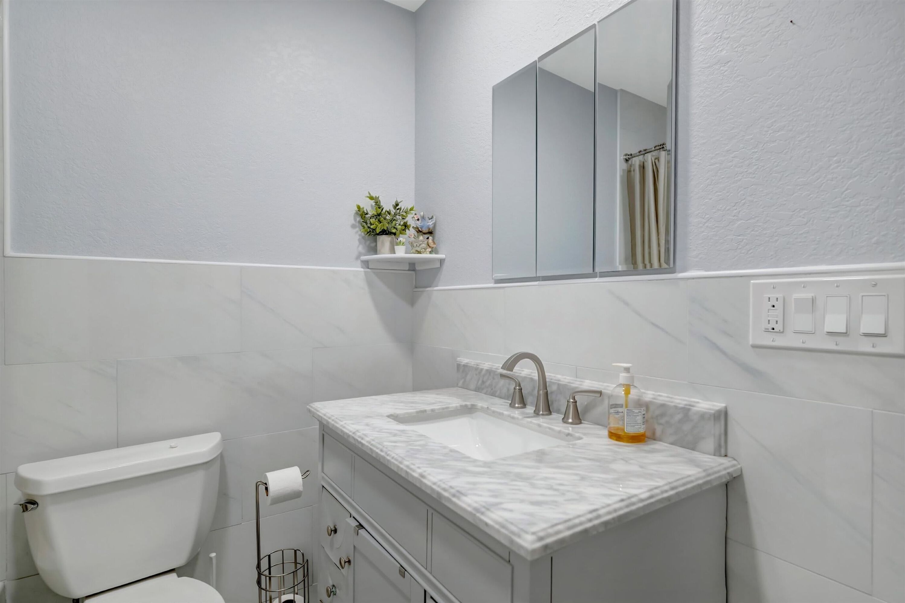 A bathroom with a toilet, a sink with a marble countertop, a mirror, and a shelf with a plant.