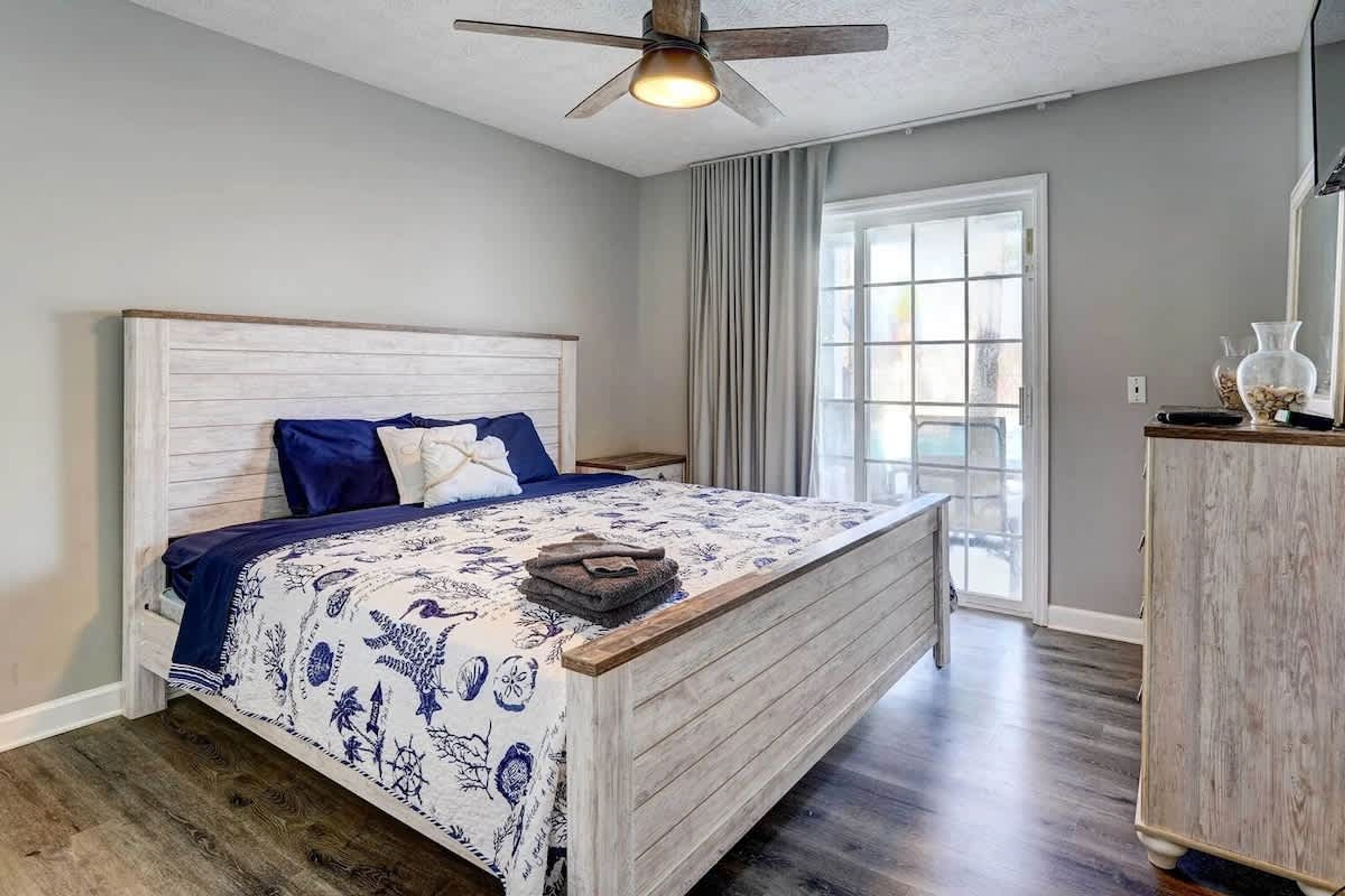 A bedroom with a wooden headboard, a bed with a patterned bedspread, a ceiling fan, a window with curtains, and a wooden dresser.