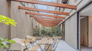 Un espace extérieur aménagé avec des sièges, protégé par un toit, comprenant une table et des chaises, entouré de plantes et offrant une vue sur un bâtiment.