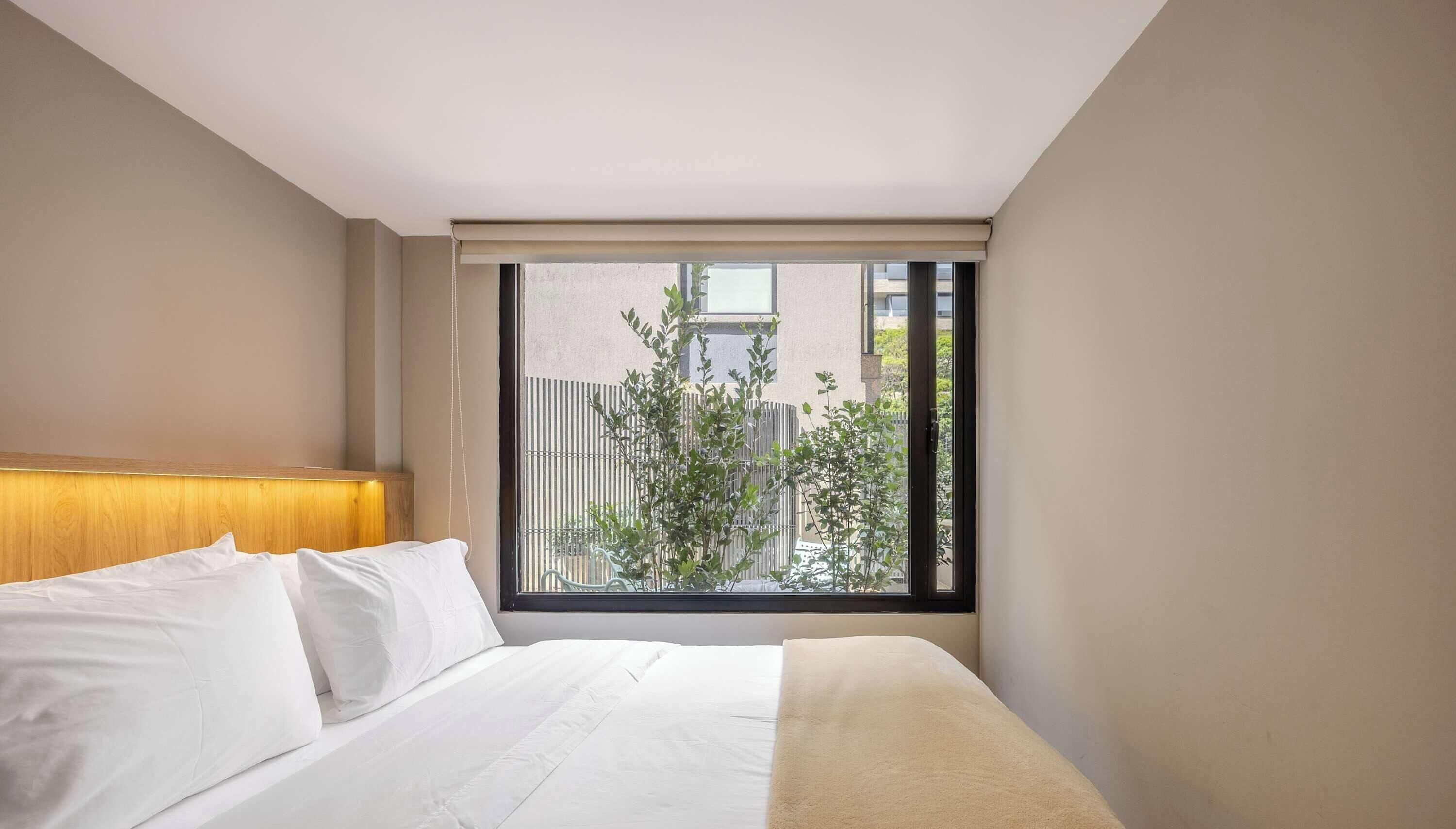 A hotel room with a bed, a headboard, a window with a view of greenery and a fence, and a ceiling with a light fixture.