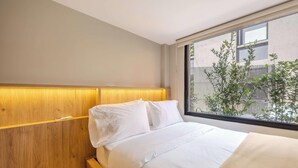 Une chambre d’hôtel avec une tête de lit en bois, une grande fenêtre donnant sur un paysage verdoyant et un lit parfaitement fait.