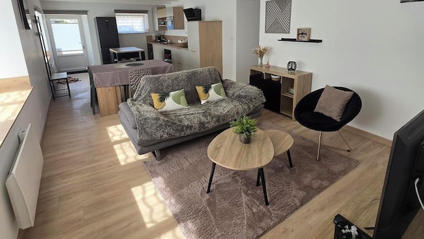 Una sala de estar moderna con un sofá, una mesa de centro y un televisor. Se observa una cocina al fondo con una zona de comedor.