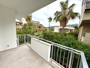 A balcony with a white railing, a tiled floor, and a view of neighboring buildings and greenery.