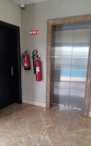 Een gang met een brandpuntteken, twee brandblussers, een deur en een metalen lift.