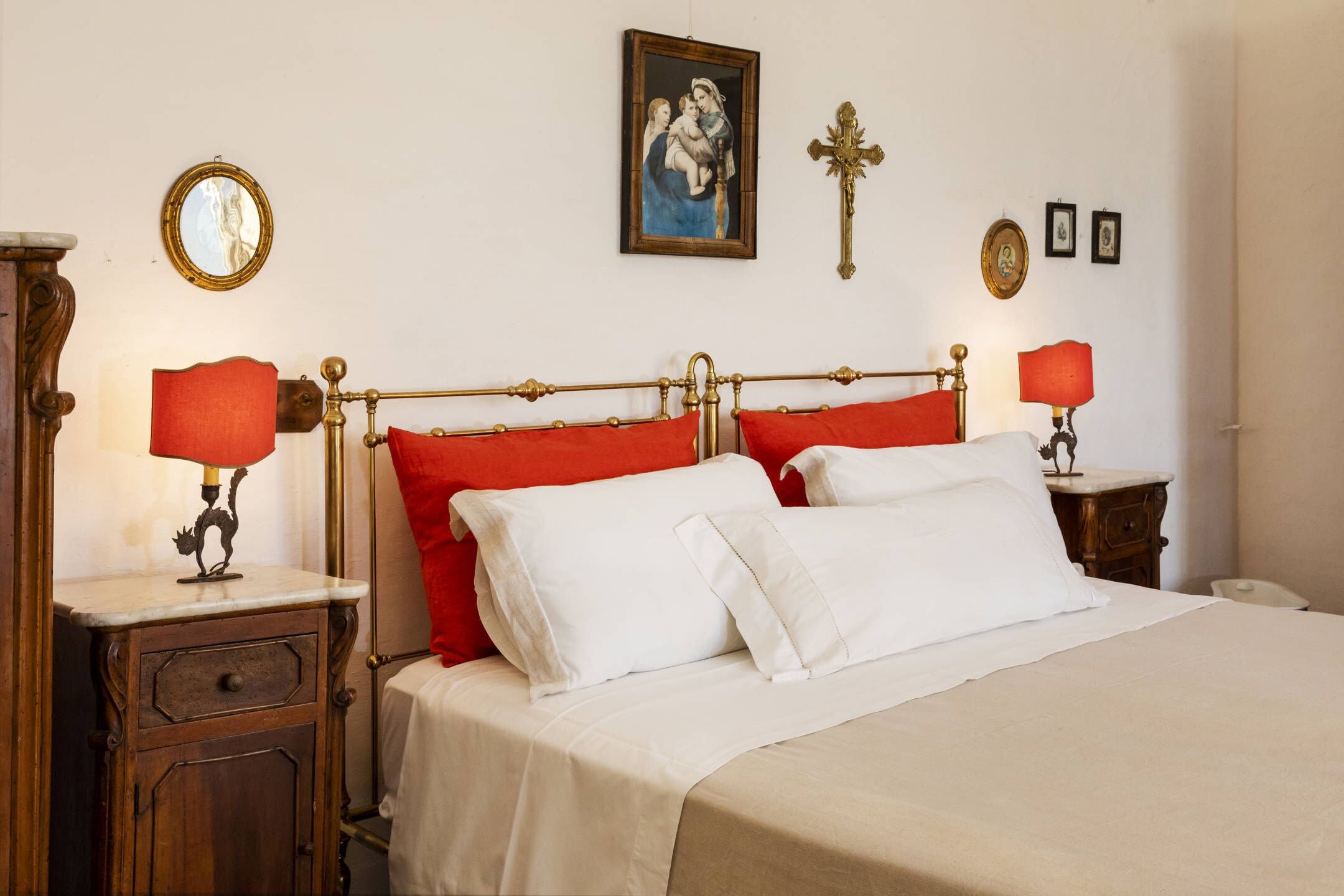 Una cama bien hecha con almohadas rojas y blancas, una mesita de noche de madera con una lámpara, una cruz colgada en la pared y un cuadro enmarcado sobre la cama.