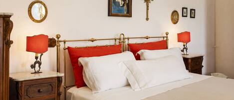 Una cama bien hecha con almohadas rojas y blancas, una mesita de noche de madera con una lámpara, una cruz colgada en la pared y un cuadro enmarcado sobre la cama.