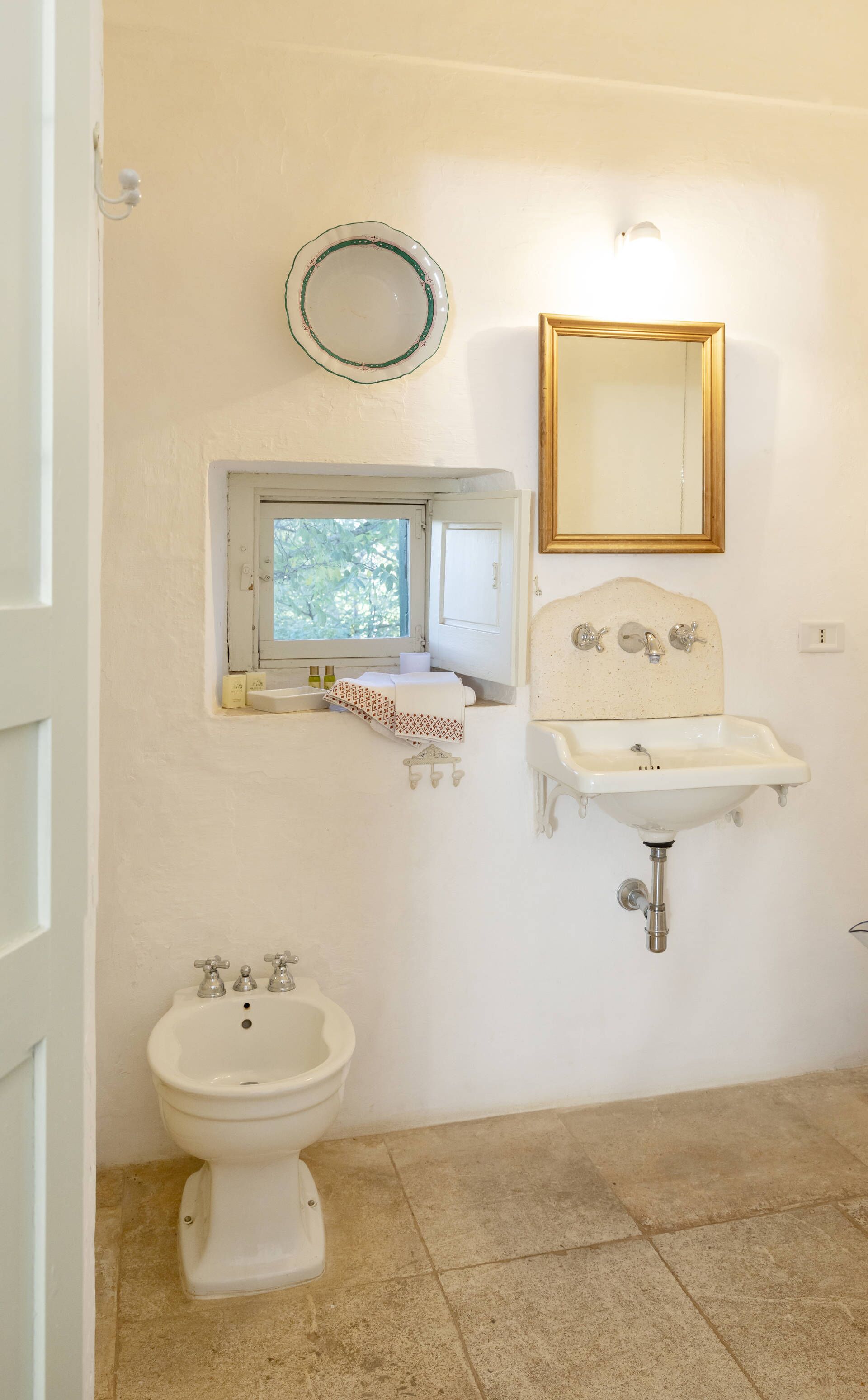 A bathroom with a toilet, bidet, sink, window, and mirror.