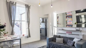 Un salon moderne avec un canapé, une table à manger et des chaises, une grande fenêtre avec des rideaux, une cuisine en arrière-plan et un miroir agrémenté d’œuvres d’art.