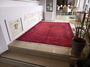 Un hall d’hôtel avec un tapis rouge à motifs, une enseigne fixée au mur portant l’inscription « ASSEMBLY HOTEL », et une plante en pot.