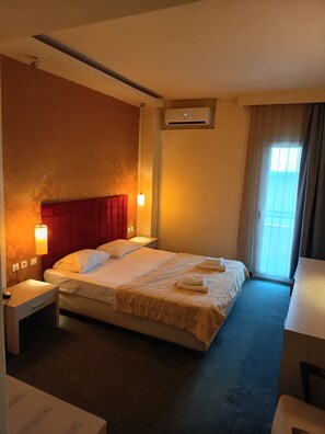 Une chambre d’hôtel comprenant un lit, des tables de chevet, un bureau, une chaise, un appareil de climatisation et une fenêtre avec des rideaux.