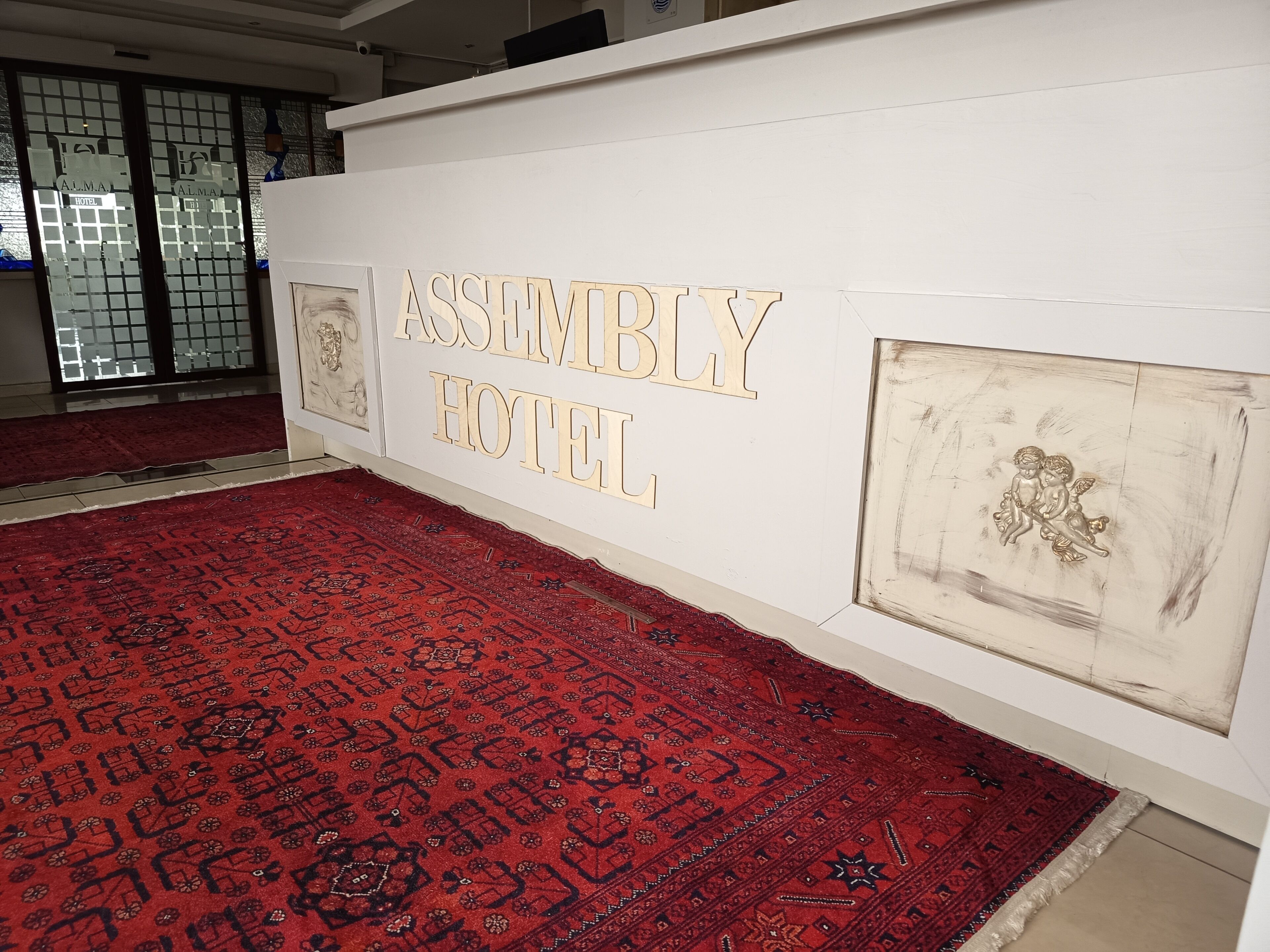 Un vestíbulo de hotel con una alfombra roja estampada, una pared con el nombre 'ASSEMBLY HOTEL' en letras grandes y dos obras de arte enmarcadas.