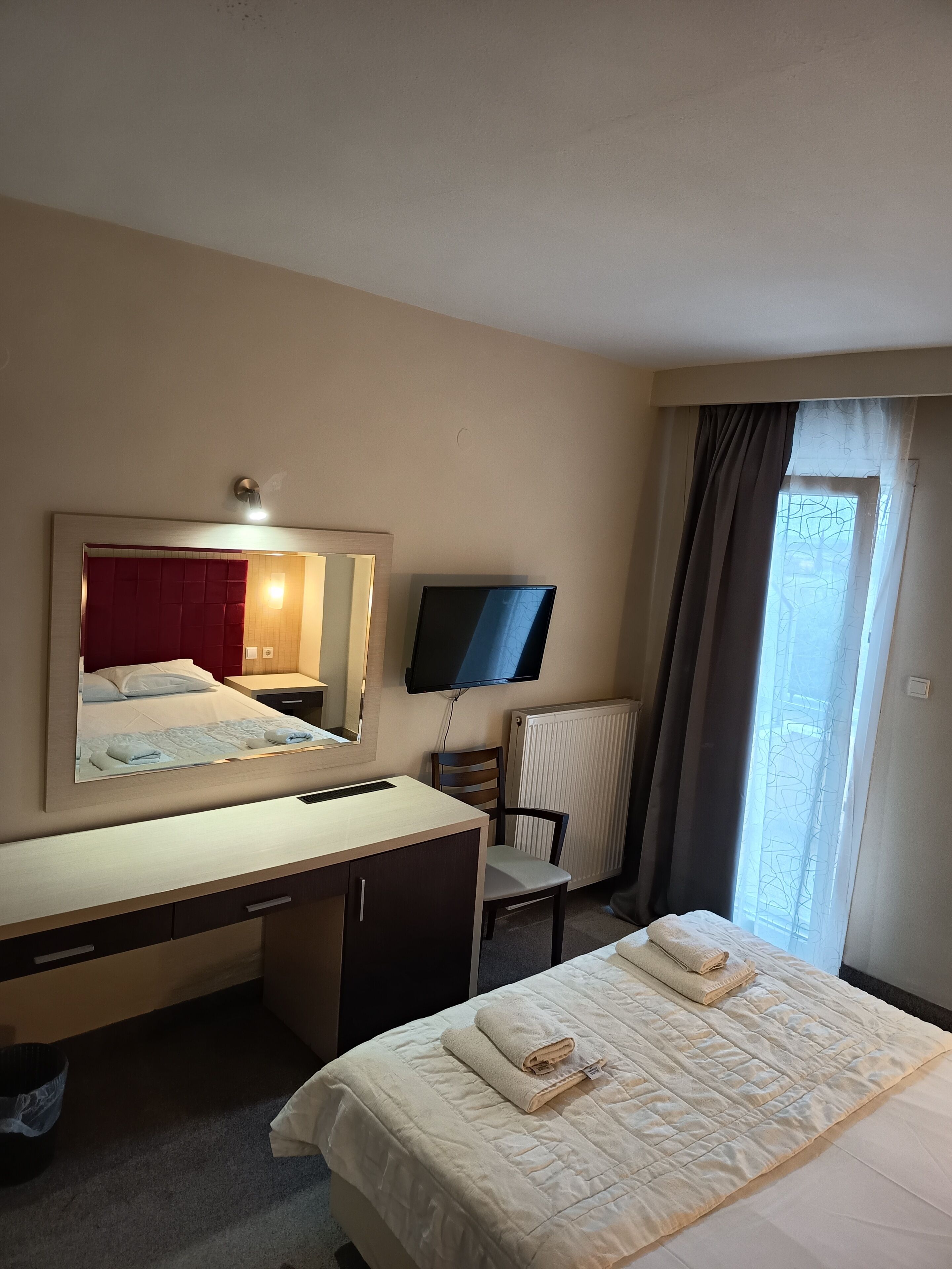 Habitación de hotel con cama, escritorio, silla, televisor, ventana con cortinas y radiador.