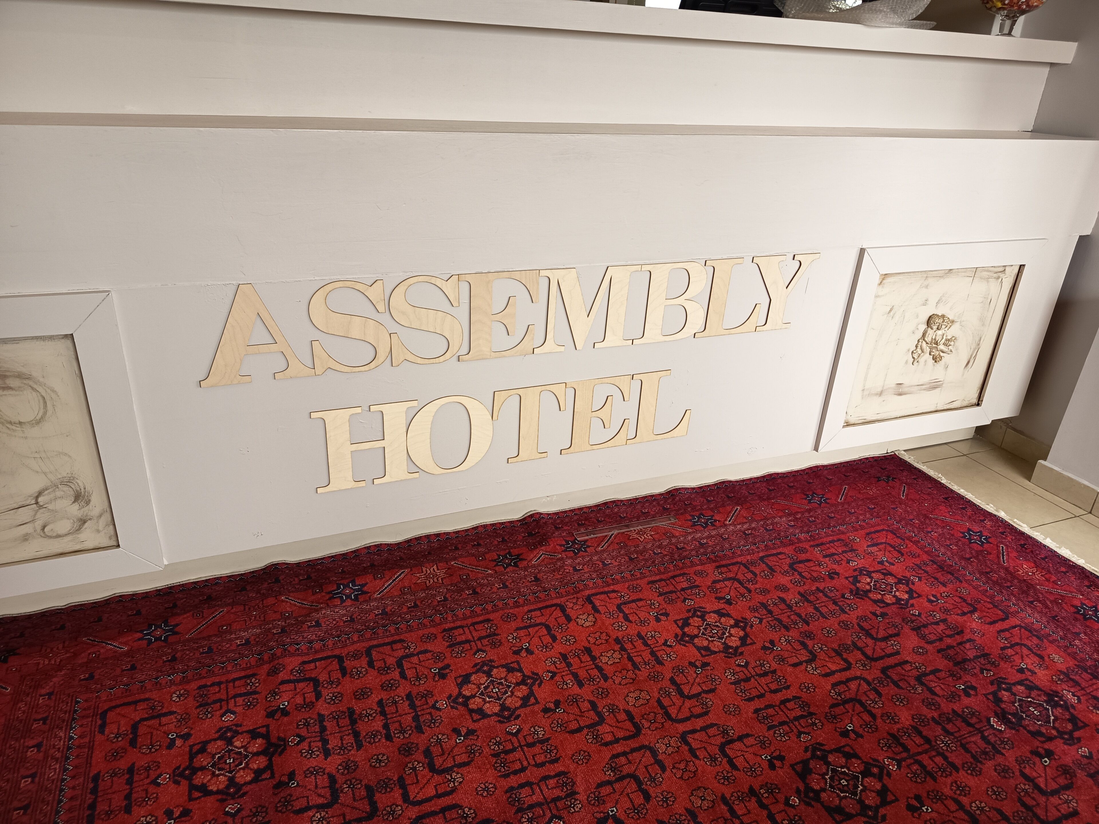 A reception desk with 'ASSEMBLY HOTEL' signage, a patterned red carpet, and a framed picture on the wall.