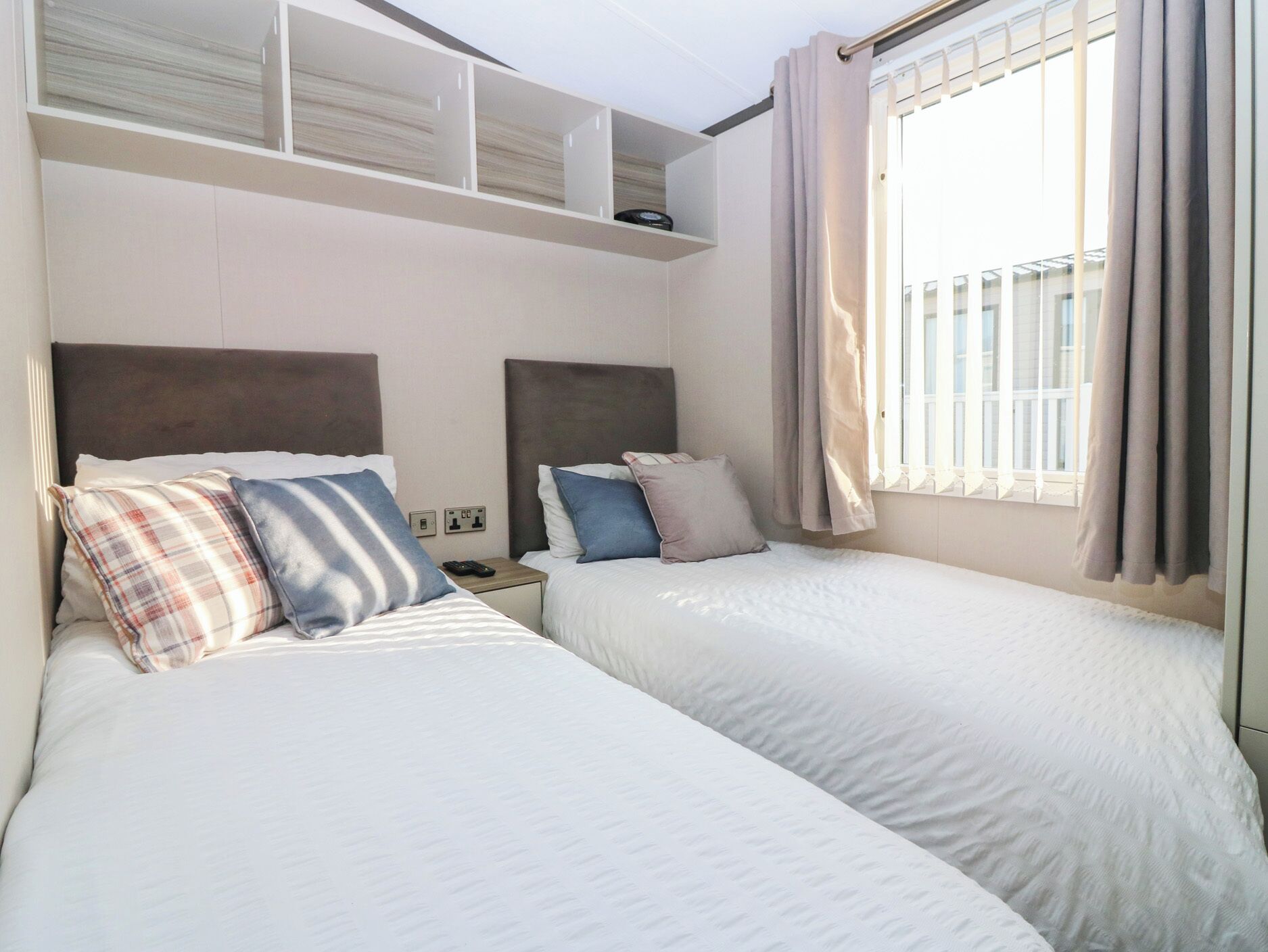 Habitación de hotel con dos camas, un estante empotrado encima de ellas, una ventana con cortinas y un enchufe en la pared.