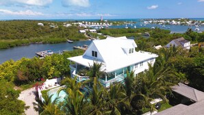 A white house with a pool, surrounded by lush greenery and a marina with boats.