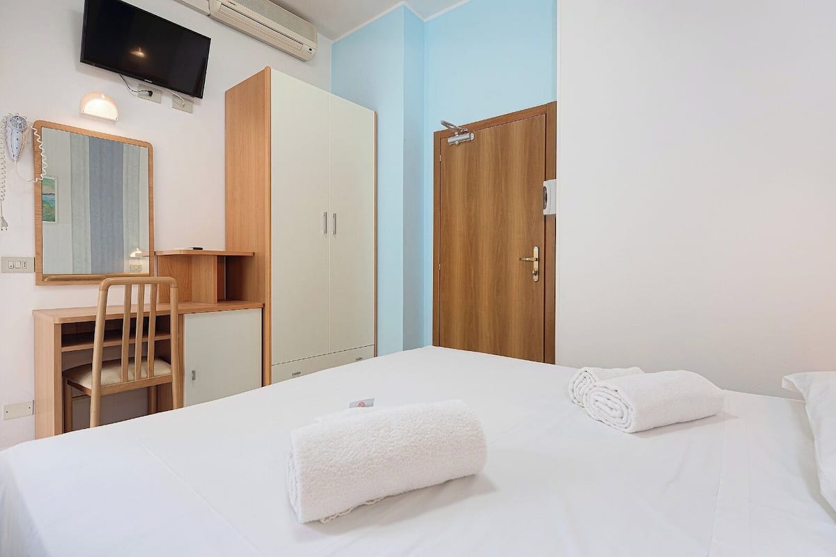 A bedroom with a bed, a desk, a chair, a wardrobe, a television, and a mirror.
