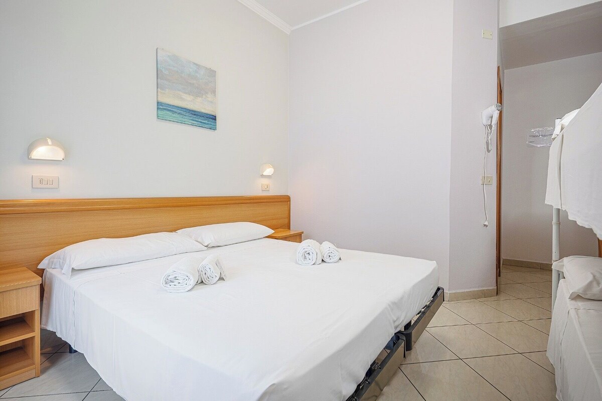 A hotel room with a bed, two bedside lamps, a painting on the wall, and a bathroom with a towel rack.