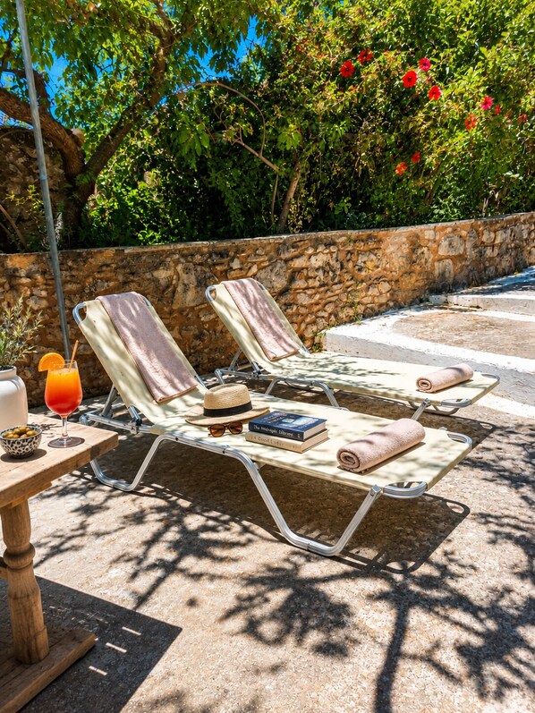 Een zonnig terras buiten met lounge fauteuils en een tafel, omringd door een stenen muur en weelderige beplanting.