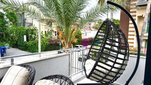 A balcony with wicker chairs, a hanging chair, and a view of a residential area with palm trees and houses. - SUNSET SUİTE 2+1 Family Apartment - 200m Walking Distance to the Beach (Kemer)
