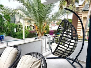 A balcony with wicker chairs, a hanging chair, and a view of a residential area with palm trees and houses.