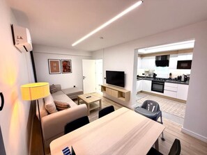 A modern apartment with a living area, dining table, sofa, TV, kitchen, and a small balcony.