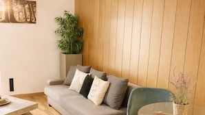 A modern living room with a grey sofa, a glass coffee table, a blue armchair, a wooden wall, and a potted plant. - Cozy Home in Nature Park (Schlangenbad)