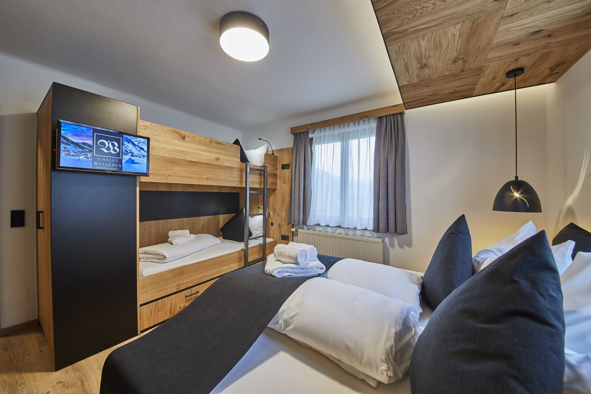 A hotel room with a bunk bed, a flat bed with pillows, a TV on the wall, a window with curtains, and a ceiling light.