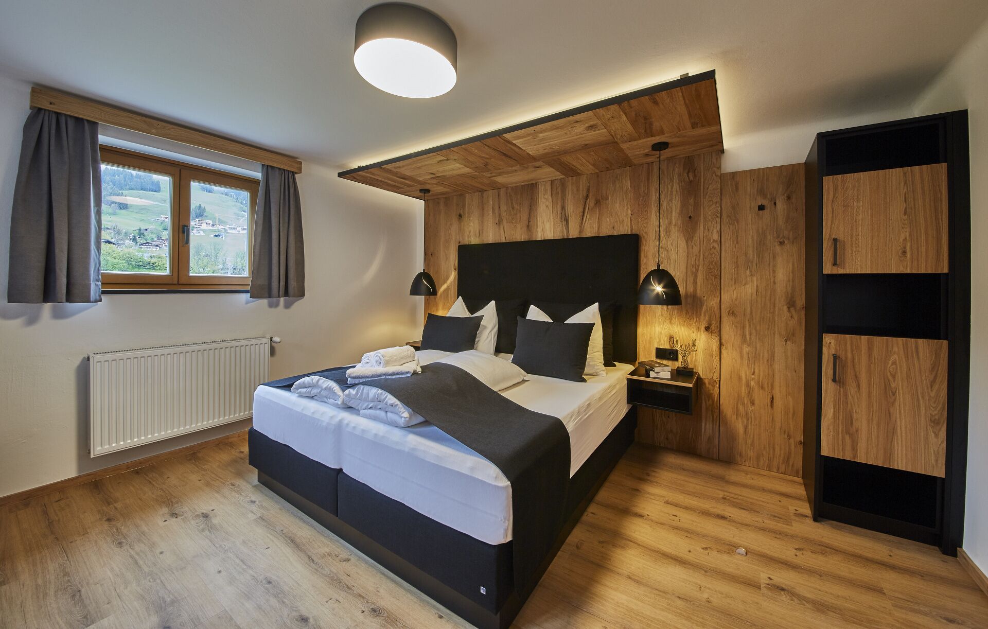 A bedroom with a bed, bedside tables, a nightstand, a radiator, a window with curtains, and a view of the outdoors.