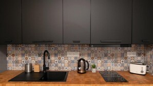 Eine Küche mit dunklen Schränken, einer gemusterten Spritzschutzleiste, einem schwarzen Spülbecken, einem Edelstahlkocher, einem Toaster und einem kleinen Topf mit Pflanze.