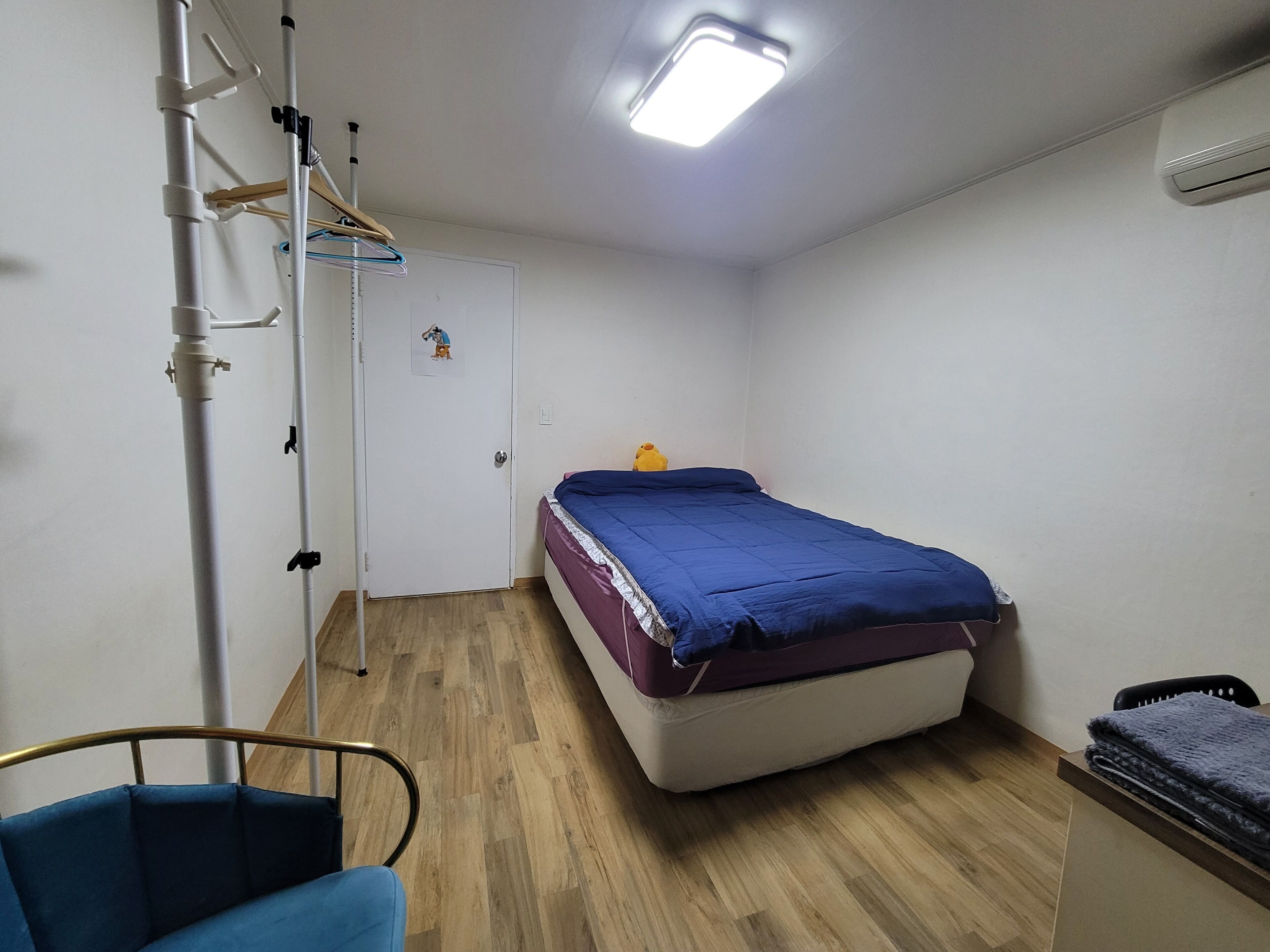 A small, single-bed room with a wooden floor, a bed with a blue blanket, a chair, a desk, and a white door with a small picture.