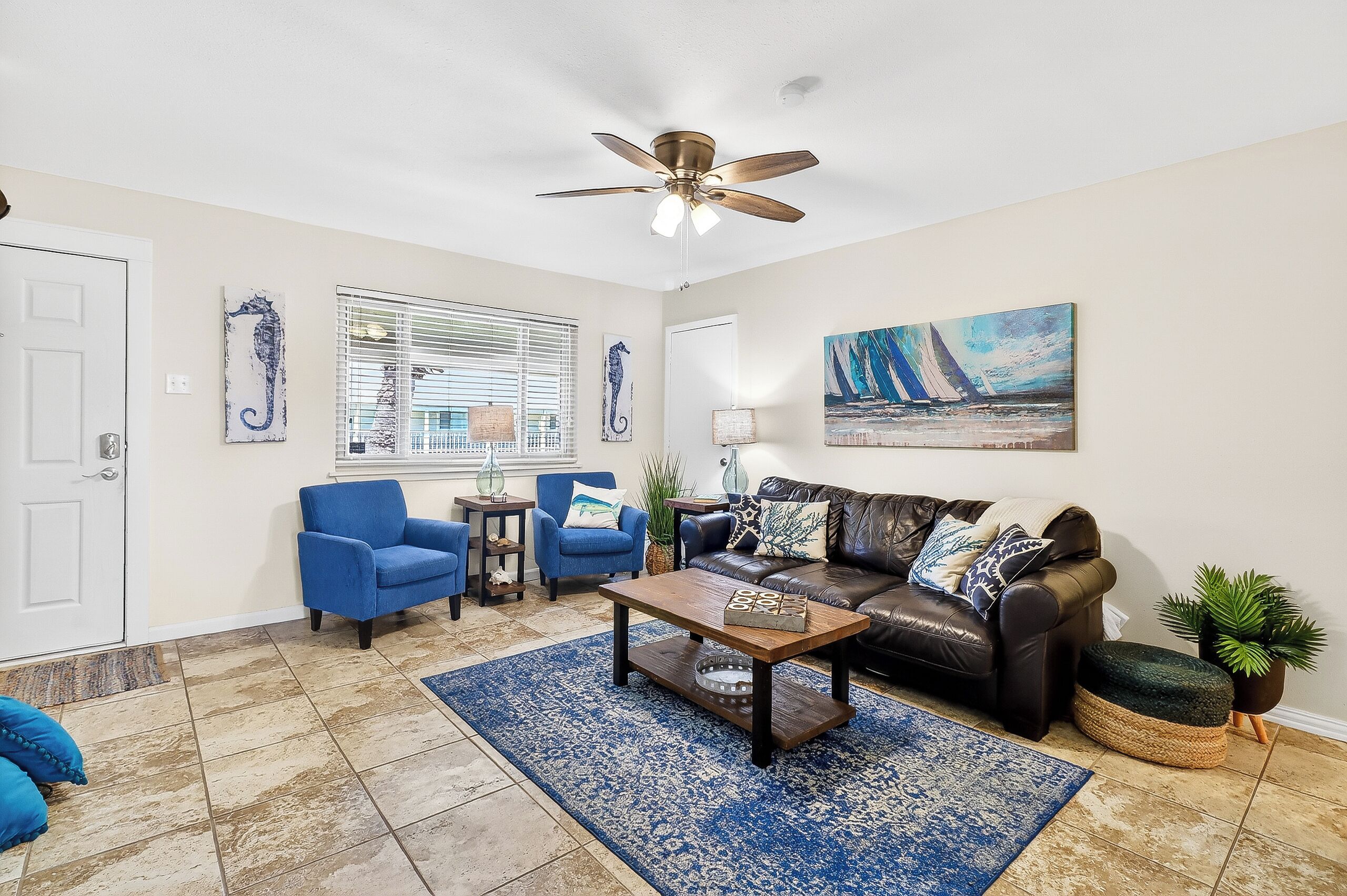 A living room with a ceiling fan, a coffee table, a sofa, two armchairs, a painting, and a window with blinds.
