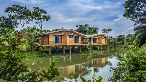 Two wooden houses on stilts over a calm body of water, surrounded by lush greenery. - Pubail Resort Club (Gazipur)