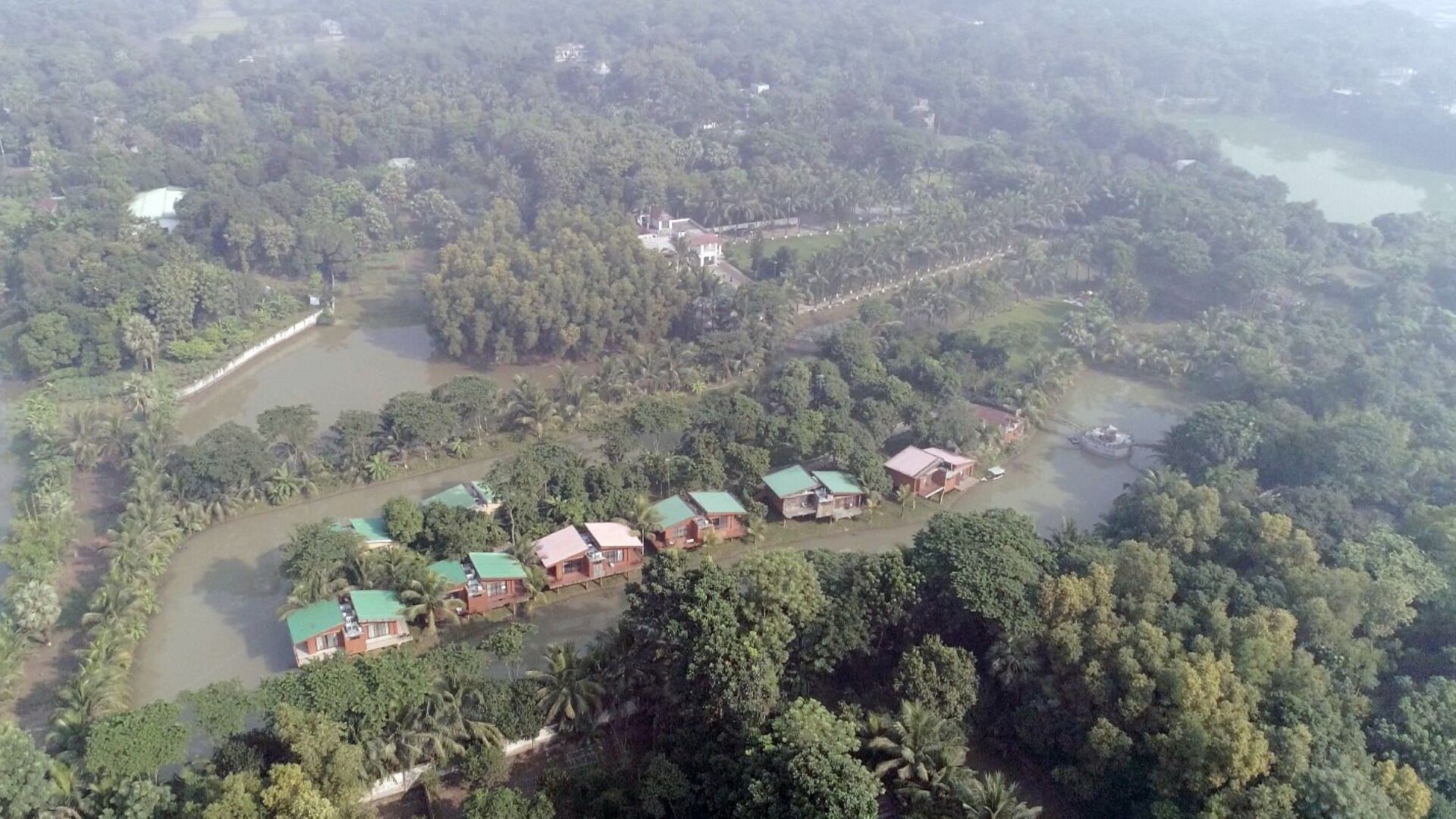 Vista aérea de un resort con varias construcciones tipo bungalow con techos verdes, rodeado de densa vegetación tropical y una masa de agua.
