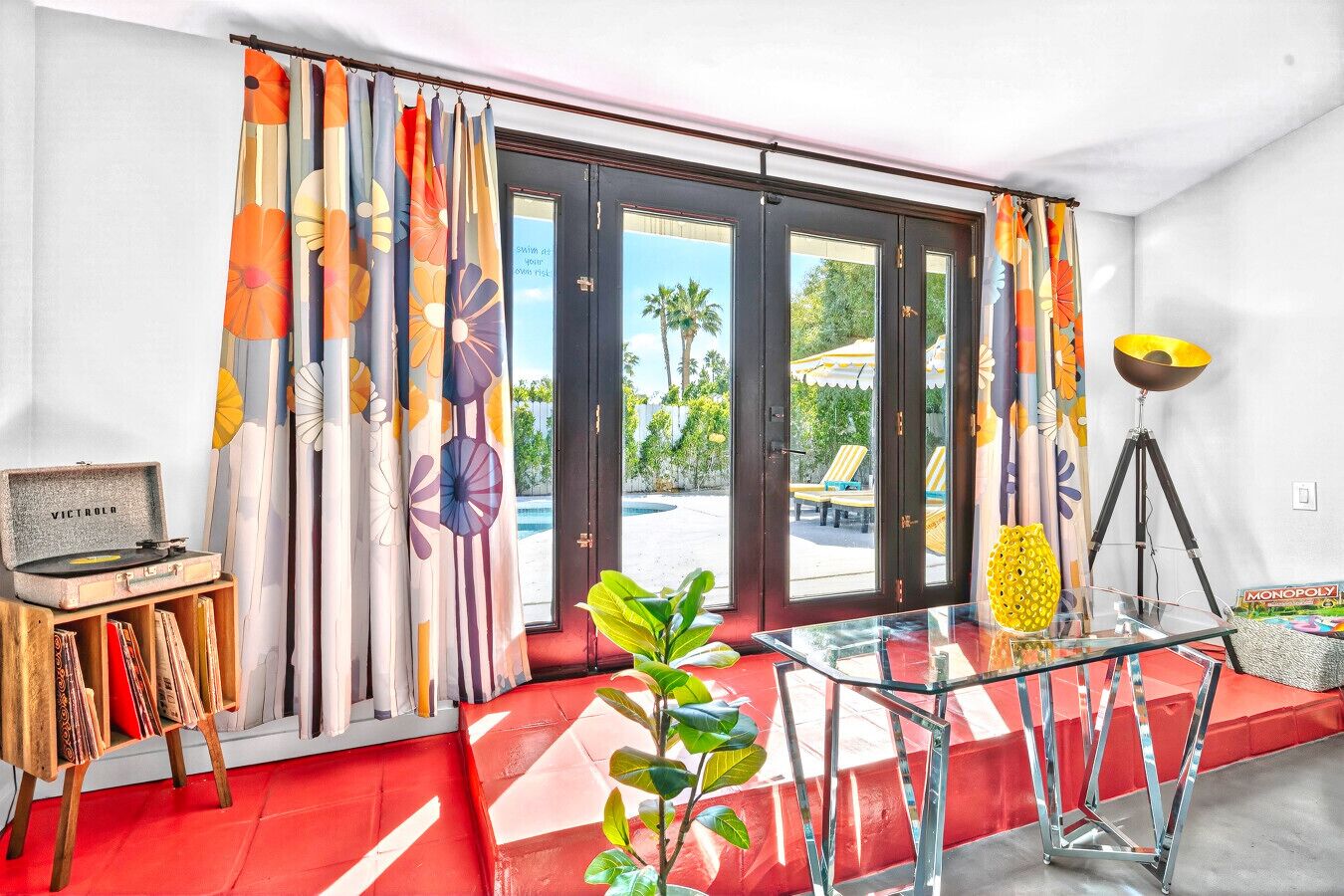 Una habitación con alfombra roja, una mesa de vidrio, un tocadiscos y vistas a una piscina y sillones de descanso al aire libre.