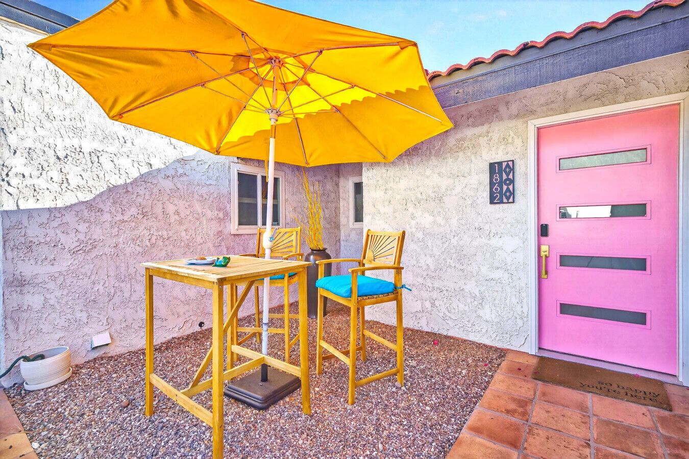 A patio with a yellow umbrella, a wooden table, two chairs, and a pink door.