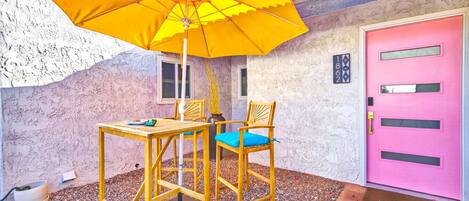 A patio with a yellow umbrella, a wooden table, two chairs, and a pink door.