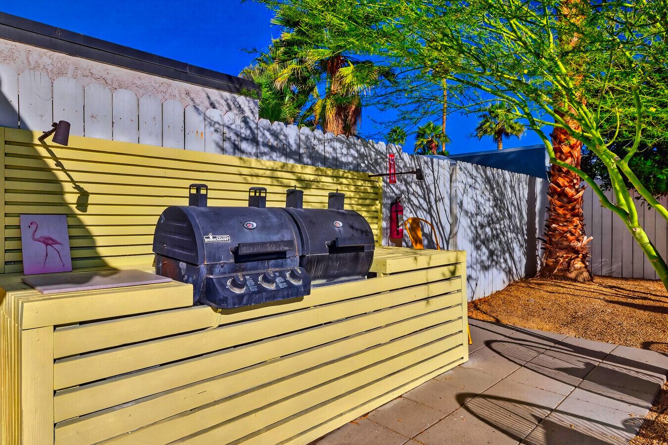 Ein Grill ist vor einem gelben Zaun, einer Holzwand und Palmen aufgebaut.
