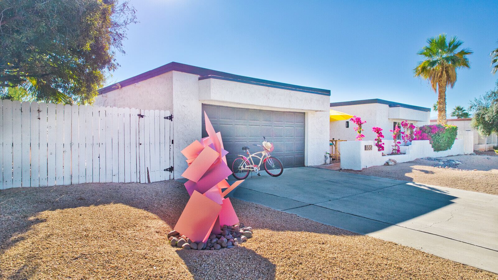 Una zona residencial con un cerco blanco, un garaje, una bicicleta y una escultura rosa.
