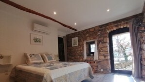 A bedroom with a stone wall, a bed with patterned bedding, a window with curtains, and an air conditioning unit. - Charming bed and breakfast in an authentic Ardèche farmhouse. (Saint-Paul-le-Jeune)