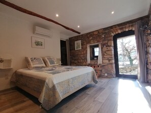 Een slaapkamer met een stenen muur, een bed met beddengoed met een patroon, een raam met gordijnen en een airconditioning.